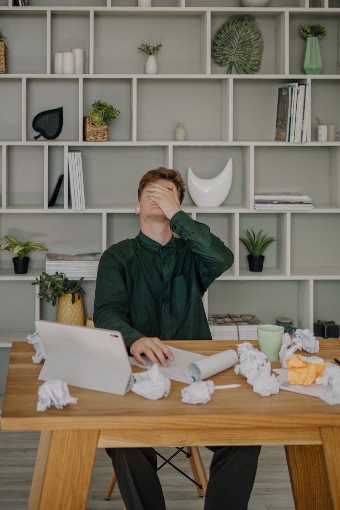 Man sitting at a desk overwhelmed with work, surrounded by crumpled papers and tablet.