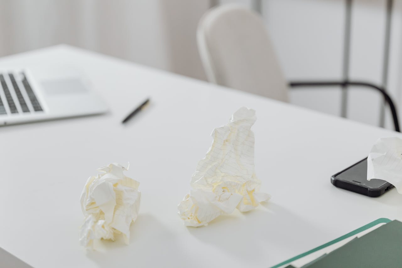 A clean white workspace featuring a laptop, phone, and crumpled papers.