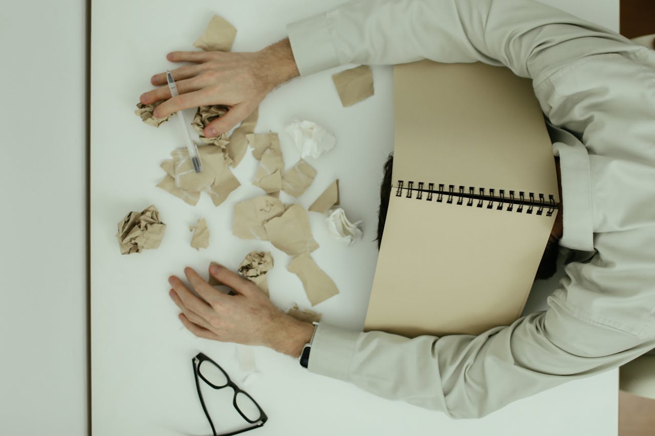 A tired adult man resting his head on a desk cluttered with crumpled papers and an open notebook.